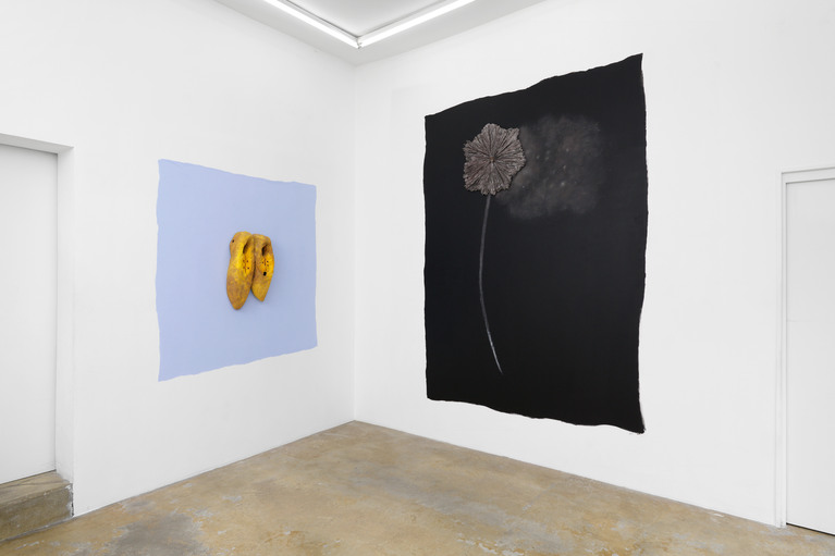 Deux oeuvres murales : une peinture bleue myosotis avec une sculpture de deux grands sabots jaunes en céramique et une peinture noire avec une fleur en céramique et de la fumée réalisée avec de la cendre